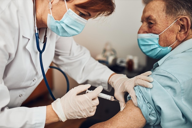 Arzt hält eine Spritze mit Impfstoff und injiziert einen älteren Patienten mit einer medizinischen Maske