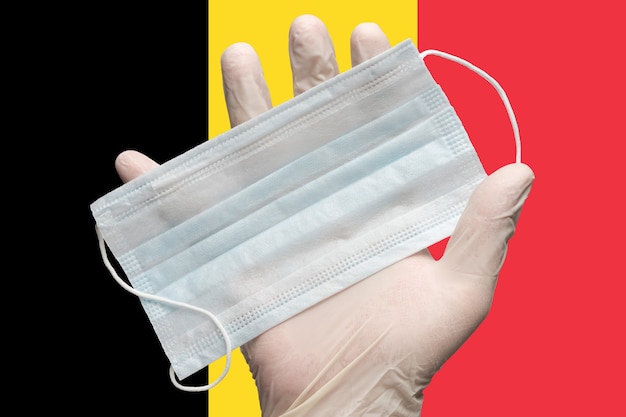 Arzt hält medizinische Gesichtsmaske in der Hand in weißen Handschuhen auf Hintergrundfarben Flagge des belgischen Konzepts