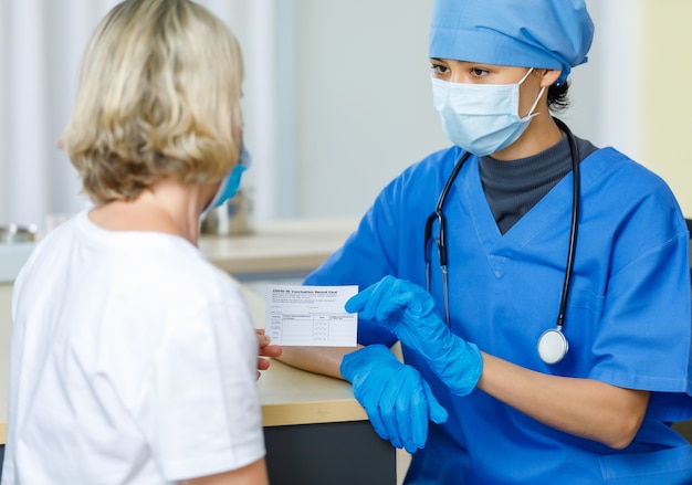 Arzt in blauer Krankenhausuniform-Gesichtsmaske und Gummihandschuhen mit Stethoskop senden und erklären das Covid-19-Impfausweiszertifikat an die kaukasische Patientin im verschwommenen Vordergrund.