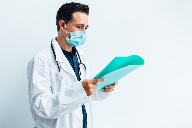 Arzt in OP-Maske, weißem Kittel, Stethoskop und blauem Ordner mit einem Bericht in der Hand.
