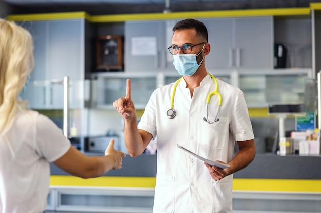 Arzt in steriler Uniform mit Gesichtsmaske beim Schieben kein Seufzen mit der Hand, weil der Patient ihm die Hand geben möchte. Andererseits hält er einen Ordner mit Testergebnissen.