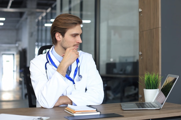 Arzt in weißer medizinischer Uniform mit Statoskop, das die persönliche Akte des Patienten auf dem Tablet liest und Daten auf den Laptop schreibt.