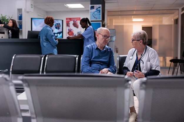 Arzt konsultiert alten Mann im Wartezimmer und spricht über Krankheitsdiagnose und -behandlung in der Krankenhauslobby. Leitender Arzt und Patient, der einen Kontrolltermin im Wartebereich der Klinik macht.