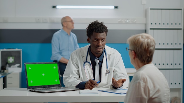 Arzt konsultiert alten Patienten und hat grünen Bildschirm auf Laptop im medizinischen Kabinett. Anzeige der Chroma-Key-Vorlage mit isoliertem Mockup-Hintergrund auf dem Schreibtisch, während der Sanitäter mit einer älteren Frau spricht