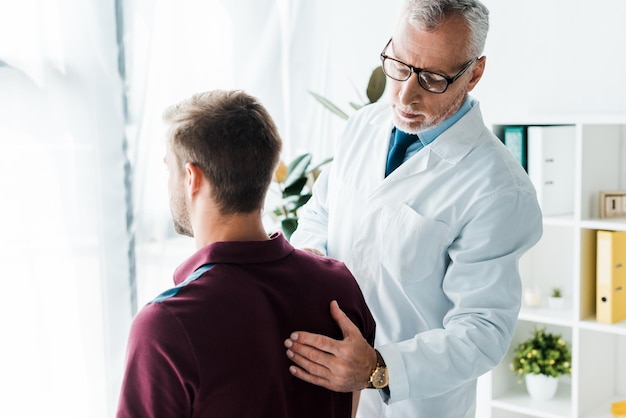 Arzt mit Brille und weißem Kittel berührt den Rücken eines Mannes in der Klinik