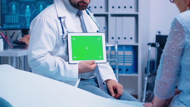 Arzt mit horizontalem Greenscreen-Chroma-Tablet vor älteren Patienten in einem privaten modernen Krankenhaus oder einer Klinik. Fertiges Chroma-Mockup für Ihre App, Ihren Text, Ihr Video oder andere digitale Assets