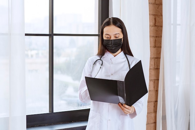Arzt mit Stethoskop und schwarzer Maske, der neben dem Fenster steht und einen schwarzen Geschichtsordner der Patienten hält, sie öffnet und liest.
