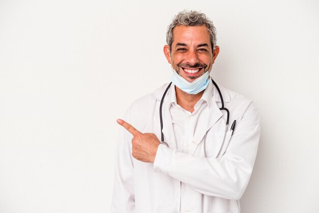 Arzt mittleren Alters, der eine Maske für Viren trägt, isoliert auf weißem Hintergrund, lächelt und zur Seite zeigt und etwas auf die Leerstelle zeigt
