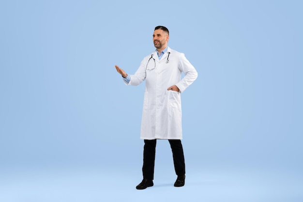 Arzt mittleren Alters zeigt Kopierraum mit der Hand in Uniform, die auf blauem Studiohintergrund in voller Länge geht