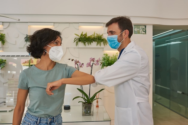 Arzt und Patient begrüßen sich, indem sie sich an den Ellbogen stoßen. Sie tragen Gesichtsmasken. Sie sind an der Rezeption der Klinik.