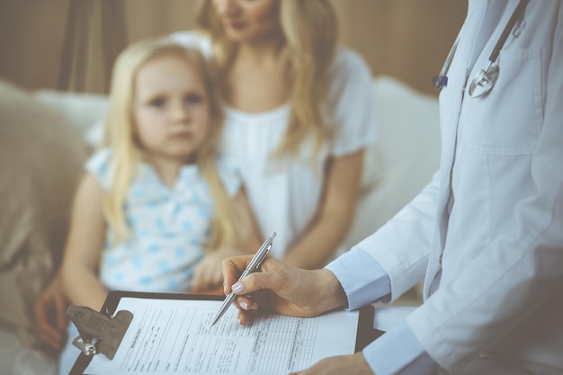 Arzt und Patient. Kinderarzt mit Klemmbrett bei der Untersuchung des kleinen Mädchens mit ihrer Mutter zu Hause. Krankes und unglückliches Kind bei der ärztlichen Untersuchung. Medizin-Konzept.