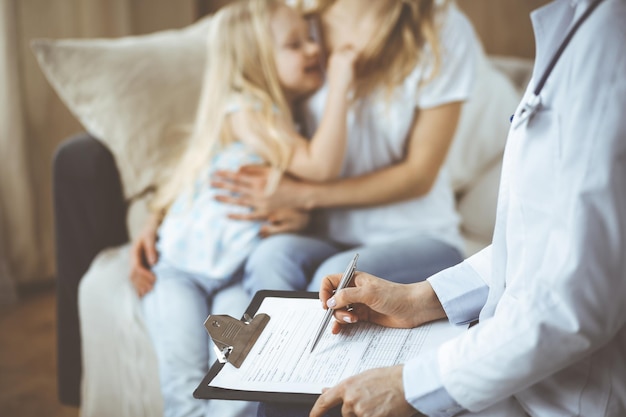 Arzt und Patient. Kinderarzt mit Klemmbrett bei der Untersuchung des kleinen Mädchens mit ihrer Mutter zu Hause. Krankes und unglückliches Kind bei der ärztlichen Untersuchung. Medizin-Konzept.