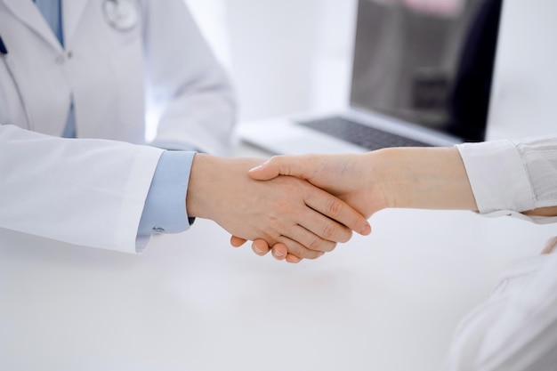 Arzt und Patient schütteln sich die Hände, während sie in der Klinik am Tisch gegenüber sitzen, nur die Hände nahe. Medizinkonzept.