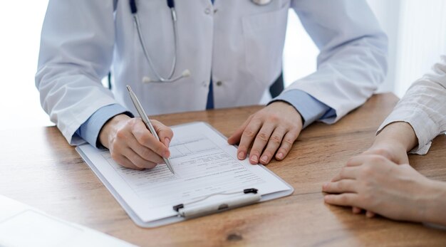 Arzt und Patient sitzen am Holztisch in der Klinik. Im Mittelpunkt stehen die Hände einer Ärztin, die ein Aktenformular ausfüllt. Medizinkonzept.