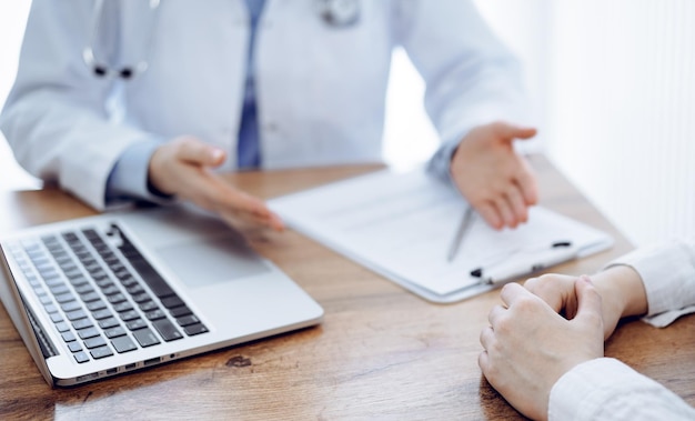 Arzt und Patient sitzen am Holztisch und besprechen etwas, während sie einen Laptop benutzen. Der Fokus liegt auf den Händen des Patienten. Medizinkonzept