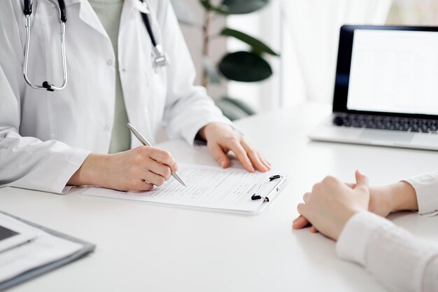 Arzt und Patient sitzen nebeneinander am weißen Schreibtisch in der Klinik. Die Ärztin hört zu und füllt ein Aufzeichnungsformular aus. Medizinkonzept.