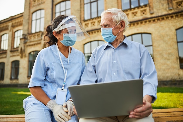 Arzt und Patient tragen Schutzmaske und sitzen mit Laptop auf Bank