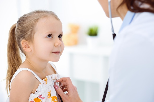 Arzt untersucht ein Kind mit Stethoskop in sonniger Klinik. Glücklich lächelnde Mädchenpatientin in hellem Farbkleid ist bei der üblichen medizinischen Untersuchung.