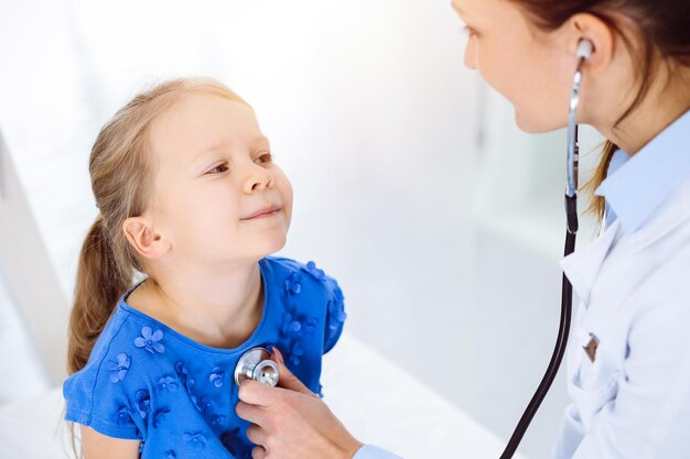 Arzt untersucht ein Kind mit Stethoskop in sonniger Klinik. Glücklich lächelnde Patientin in blauem Kleid ist bei der üblichen ärztlichen Untersuchung.