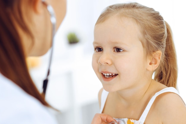 Arzt untersucht ein kleines Mädchen mit einem Stethoskop. Glücklich lächelnder Kinderpatient bei der üblichen ärztlichen Untersuchung. Medizin- und Gesundheitskonzepte.