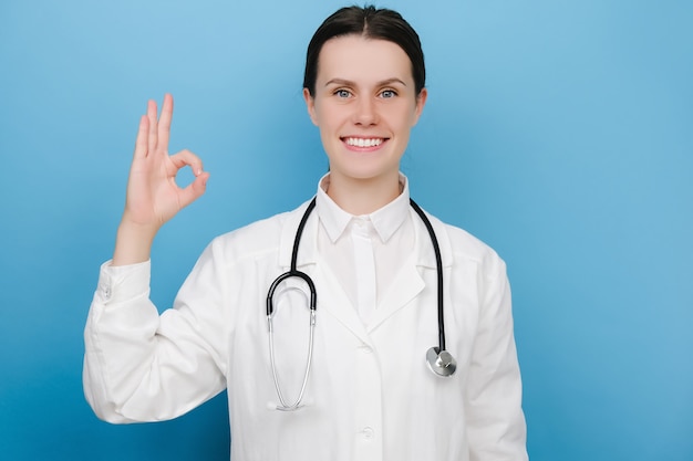 Arzt zeigt OK. Positive lächelnde Ärztin auf blauem Studiohintergrund mit Blick auf die Kamera und mit Gestenhand, die OK-Zeichen zeigt, trägt weiße Uniform und Stethoskop. Menschen Emotion Lifestyle-Konzept