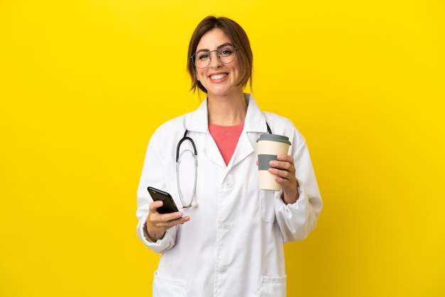 Arztfrau isoliert auf gelbem Hintergrund mit Kaffee zum Mitnehmen und einem Handy