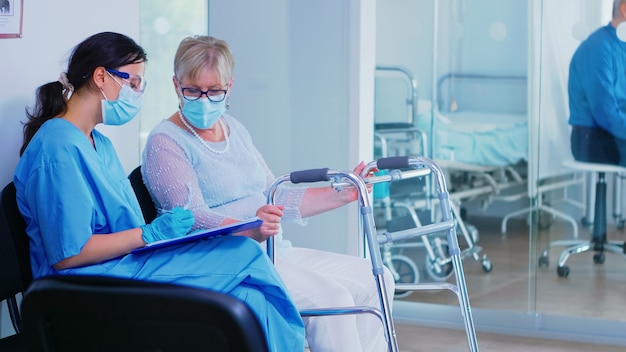 Arzthelferin mit Gesichtsmaske gegen Coronavirus im Wartezimmer des Krankenhauses, die einer ungültigen älteren Frau mit Gehrahmen hilft, Dokumente zu füllen. Patient betritt Krankenhausflur.