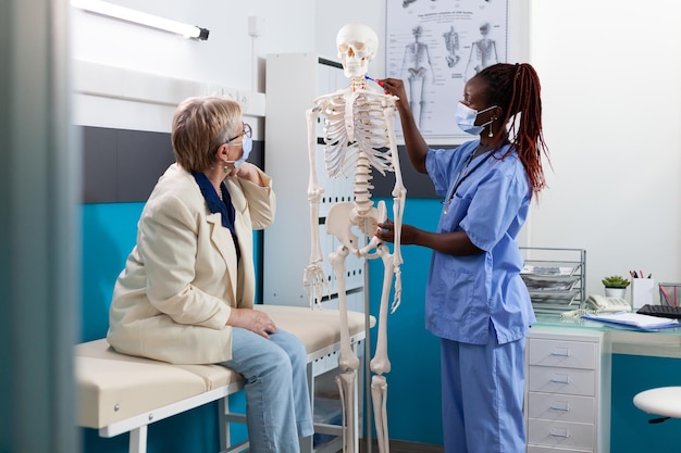 Arztkrankenschwester mit schützender Gesichtsmaske, die die anatomische Körperstruktur mit medizinischem Skelett erklärt und Rückenschmerzen mit pensionierter alter Frau während der Ernennung im Krankenhausbüro diskutiert. Medizinkonzept