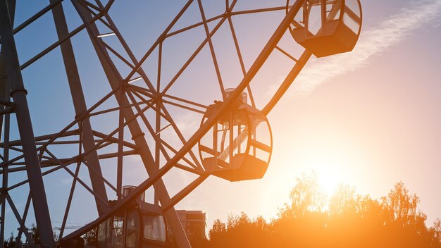 As cabines vazias de uma grande roda gigante de metal em funcionamento giram em um parque local contra o céu sobre as silhuetas das árvores à noite, após o pôr do sol