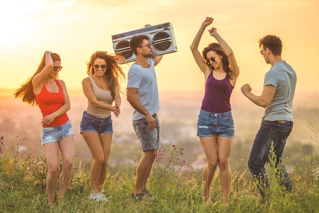 As cinco pessoas felizes dançando com um aparelho de som no fundo do pôr do sol