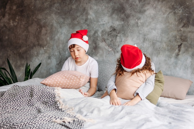 As crianças bonitos tween em chapéus de papai noel não querem acordar na cama com travesseiro no quarto loft, na manhã de natal