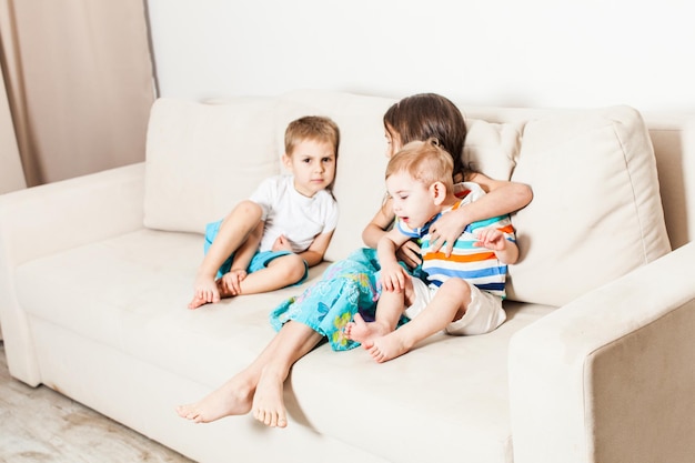 As crianças ficam entediadas durante uma sessão de fotos em casa. a irmã e dois irmãos mais novos estão sentados em um sofá branco.