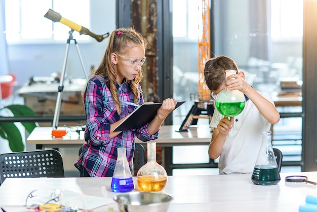 As crianças registram os resultados dos experimentos no caderno. Dois jovens alunos caucasianos inteligentes em óculos de proteção fazendo experimento com líquido verde na taça e gelo seco.