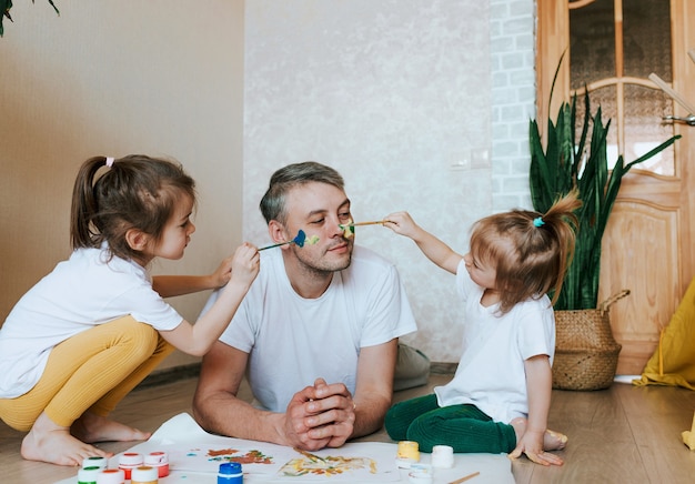 As crianças se divertem com o pai, as meninas desenham na pele do rosto de um homem com tintas coloridas, criatividade e imaginação, dia dos pais e o conceito de família