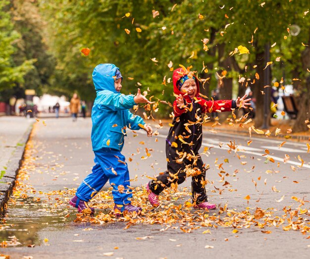 As crianças sorridentes estão brincando com folhas caídas durante a caminhada de outono Menino e menina em roupas quentes vomitam folhas