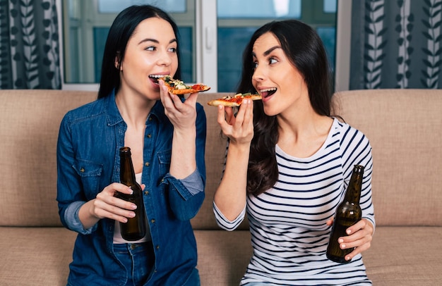 As duas melhores amigas felizes em casa no sofá assistem filmes e comem pizza vegana e bebem cerveja