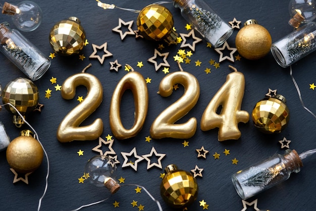 As figuras douradas 2024 feitas de velas em um fundo de ardósia de pedra preta são decoradas com uma decoração festiva de estrelas lantejoulas ramos de abeto bolas e guirlandas Cartão de feliz ano novo