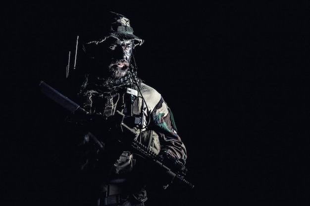 As forças especiais barbudo dos Estados Unidos em estúdio de uniformes de camuflagem tiro fundo preto com metade do comprimento. Segurando armas, usando chapéu de selva, lenço Shemagh, ele está pronto para matar. Retroiluminado