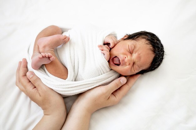 As mãos da mãe de uma mulher de pele branca estão balançando um bebê afro-americano recém-nascido em uma fralda acalmando-o antes de ir para a cama uma criança pequena está deitada na cama no quarto em close-up