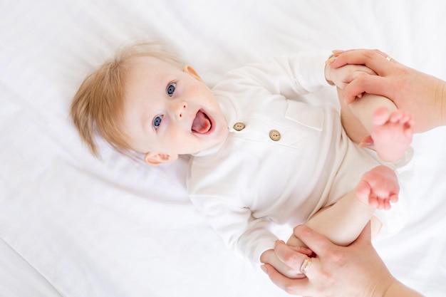 As mãos da mãe estão segurando um bebê de 6 meses, um menino loiro de olhos azuis em uma cama branca em um quarto claro em uma roupa de algodão branco o conceito de artigos infantis