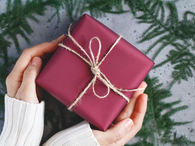 As mãos da menina em um suéter quente branco de inverno são seguradas uma caixa de presente de Natal vermelha é amarrada de barbante entre a decoração de inverno Vista superior plana