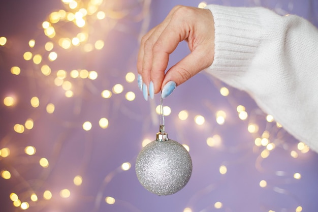 As mãos da mulher com unhas segurando uma bola de brinquedo de natal em um fundo roxo festivo bokeh desfocado