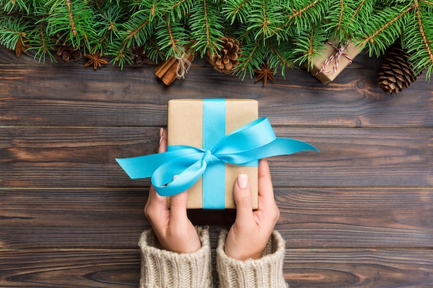 As mãos da mulher dão o presente handmade do Natal envolvido no papel com fita azul. Caixa de presente de férias na mesa de madeira escura, vista superior
