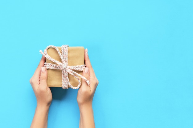 As mãos da mulher dão o Valentim envolvido ou o outro presente feito a mão do feriado no papel com fita azul. Caixa de presente, decoração de presente na mesa, vista superior com espaço de cópia