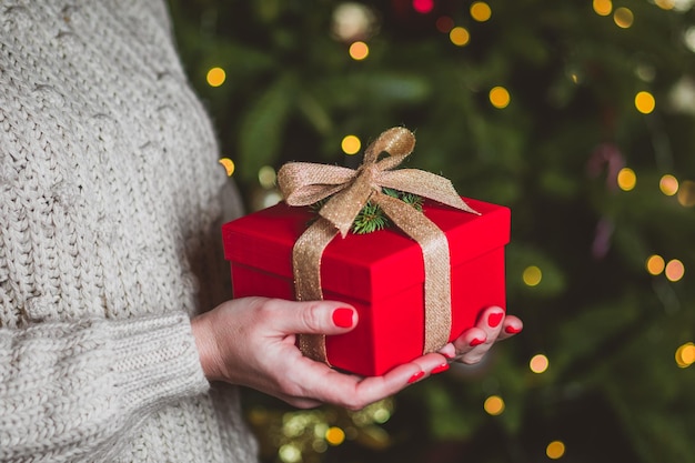 As mãos da mulher estão segurando uma caixa vermelha festiva no fundo da árvore de natal o presente de natal é embrulhado em uma fita dourada e decorado com um galho de abeto