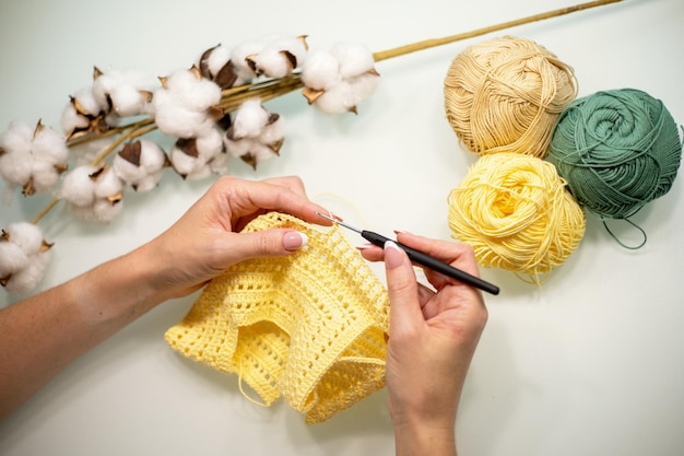 As mãos da mulher estão tricotando roupas de bebê de fio amarelo em uma vista de mesa branca