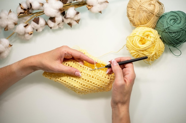 As mãos da mulher estão tricotando roupas de bebê de fio amarelo em uma vista de mesa branca