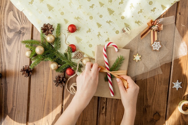 As mãos da mulher que envolvem o presente do Natal, fim acima. Presentes de Natal despreparados em madeira com elementos de decoração e itens, vista superior. Embalagem de Natal ou ano novo DIY.