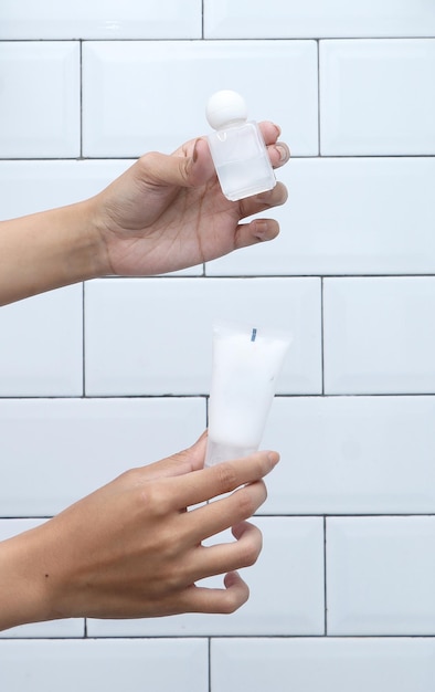 As mãos da mulher seguram recipientes cosméticos em branco para cuidados com a pele em azulejo branco minimalista no fundo.