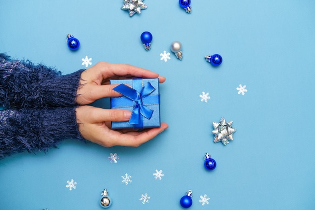 As mãos da mulher segurando uma caixa de presente presente amarrada com um laço de fita sobre fundo azul ao redor da árvore de Natal ...